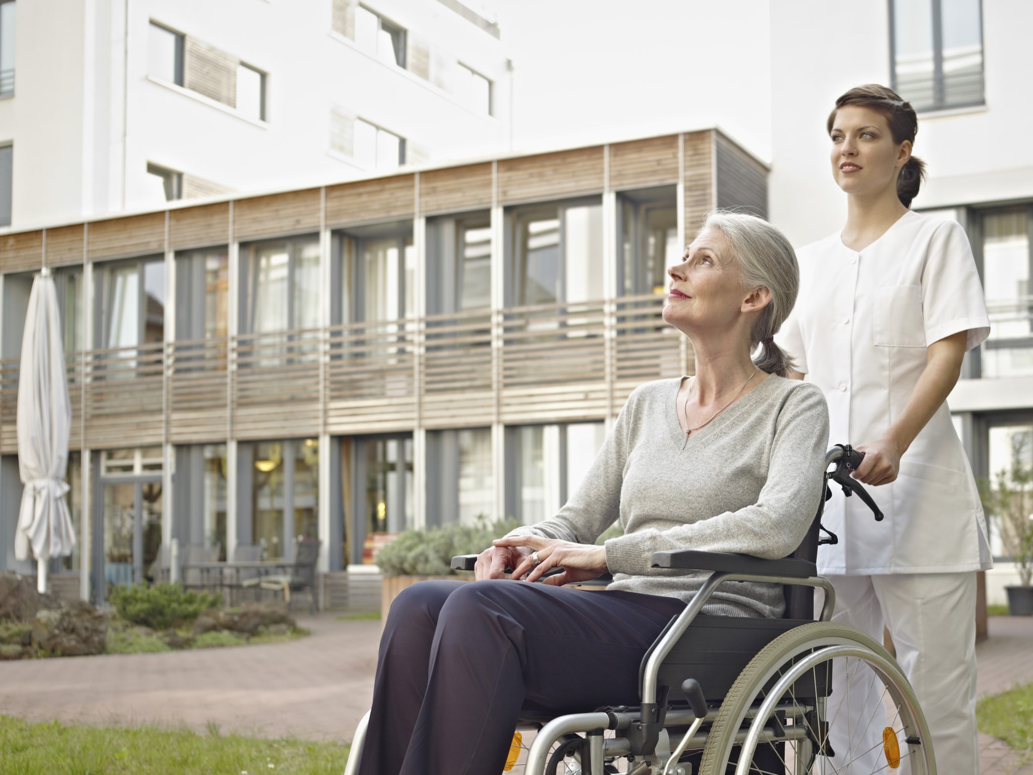 Germany, Cologne, Caretaker pushing senior women in wheelchair Seniorin wird von einer Frau in weiß vor einem mehrstöckigen Gebäude im Rollstuhl geschoben