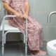 Frau mit geblümten rosa Nachthemd sitzt im Badezimmer. Zusehen sind Alltagshilfen wie eine Toilettenerhöhung, ein Duschstuhl und ein Haltegriff.