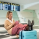 Seniorin am Flughafen. Lesend hat sie die beine auf ihren blauen Reisekoffer gelegt